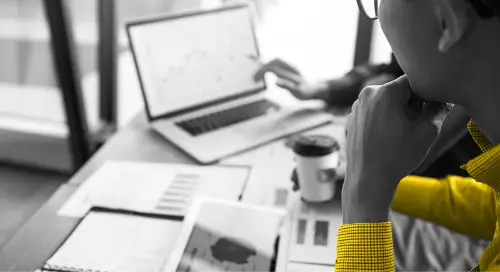 Person in a yellow shirt holding a cup of coffee and looking at a laptop