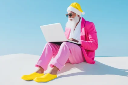 Santa wearing pink and yellow on a beach with a laptop.