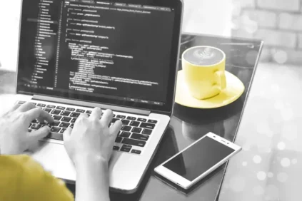 Hands on a laptop with yellow steaming mug and cell phone nearby